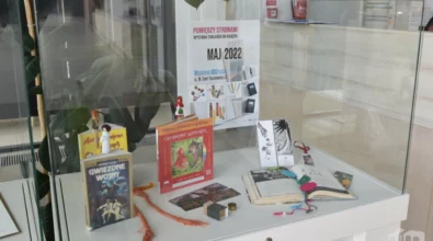 W szklanej gablocie zaprezentowane są książki, zakładki oraz drobne przedmioty inspirowane literaturą, ułożone wokół plakatu promującego wydarzenie kulturalne. Zdjęcie przedstawia fragment wystawy „Pomiędzy stronami” prezentowanej w Mediatece 800-lecia.