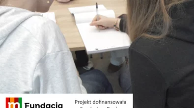 Zdjęcie przedstawia młodych uczestników siedzących przy stole i zapisujących notatki w zeszycie podczas zajęć edukacyjnych, z widocznym fragmentem grafiki informującej, że projekt został dofinansowany przez Fundację mBanku.