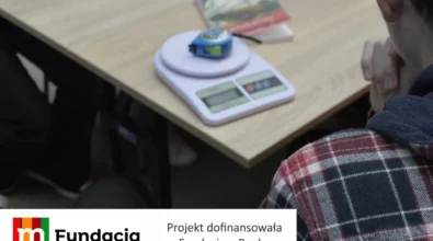 Zdjęcie przedstawia fragment zajęć edukacyjnych, na którym na stole widoczna jest waga elektroniczna z położoną na niej miarką, a obok siedzi uczestnik obserwujący ćwiczenie, z informacją, że projekt został dofinansowany przez Fundację mBanku.