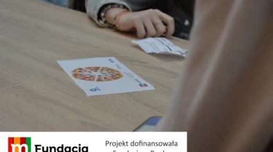 Zdjęcie przedstawia fragment warsztatów edukacyjnych, podczas których uczestnicy analizują kartę z ilustracją podzielonej pizzy leżącą na stole w ramach zajęć realizowanych w projekcie dofinansowanym przez Fundację mBanku.