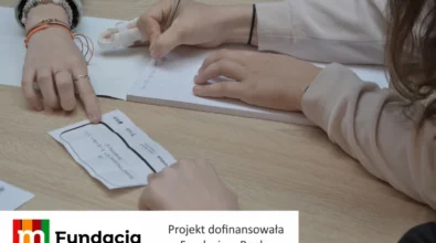 Zdjęcie przedstawia uczestników warsztatów edukacyjnych wykonujących zadania matematyczne przy stole, zapisujących obliczenia w zeszycie i analizujących karty z poleceniami w ramach projektu dofinansowanego przez Fundację mBanku.