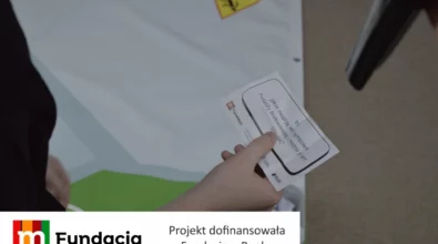 Zdjęcie przedstawia uczestnika warsztatów trzymającego kartę z pytaniem edukacyjnym podczas pracy zespołowej, z widoczną informacją, że projekt został dofinansowany przez Fundację mBanku.