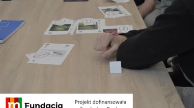 Zdjęcie przedstawia uczestników warsztatów pracujących przy stole z kartami edukacyjnymi, na których znajdują się ilustracje i zadania, podczas zajęć realizowanych w ramach projektu dofinansowanego przez Fundację mBanku.