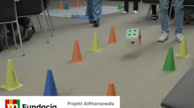 Zdjęcie przedstawia fragment warsztatów edukacyjnych, podczas których uczestnicy biorą udział w aktywności ruchowej z wykorzystaniem kolorowych pachołków i dużej kostki do gry, ustawionych na podłodze między krzesłami, w ramach projektu dofinansowanego przez Fundację mBanku.