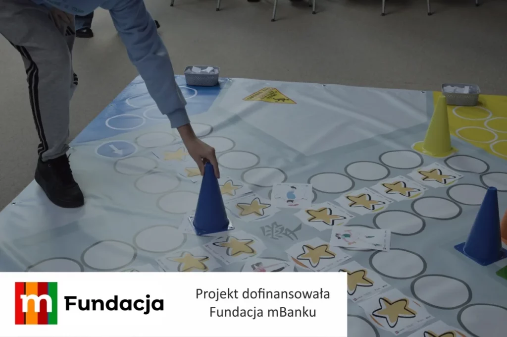 Zdjęcie przedstawia uczestnika warsztatów edukacyjnych wykonującego zadanie na dużej planszy podłogowej z wykorzystaniem kart, pachołków i elementów gry ruchowej, realizowanej w ramach projektu dofinansowanego przez Fundację mBanku.