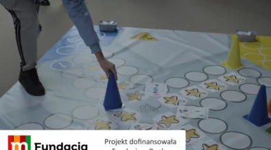 Zdjęcie przedstawia uczestnika warsztatów edukacyjnych wykonującego zadanie na dużej planszy podłogowej z wykorzystaniem kart, pachołków i elementów gry ruchowej, realizowanej w ramach projektu dofinansowanego przez Fundację mBanku.