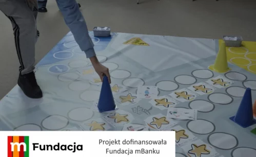 Zdjęcie przedstawia uczestnika warsztatów edukacyjnych wykonującego zadanie na dużej planszy podłogowej z wykorzystaniem kart, pachołków i elementów gry ruchowej, realizowanej w ramach projektu dofinansowanego przez Fundację mBanku.
