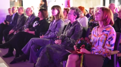 Publiczność siedzi w rzędach krzeseł i z uwagą obserwuje wydarzenie odbywające się na scenie. Na pierwszym planie kilka osób siedzi obok siebie, a jedna z kobiet trzyma w dłoniach bukiet kwiatów. Sala oświetlona jest fioletowym światłem scenicznym, które nadaje wydarzeniu uroczysty charakter.