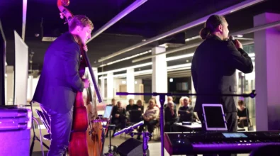 Dwóch muzyków występuje na scenie podczas koncertu. Jeden z nich śpiewa do mikrofonu, a drugi gra na kontrabasie. W tle widać siedzącą publiczność oraz oświetlenie sceniczne w odcieniach fioletu, a na pierwszym planie znajdują się instrumenty muzyczne i sprzęt nagłośnieniowy.