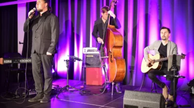 Wokalista śpiewa do mikrofonu podczas koncertu odbywającego się na scenie w Mediatece. Obok niego muzyk gra na kontrabasie, a drugi akompaniuje na gitarze akustycznej. Scena oświetlona jest fioletowym światłem, a w tle widoczne są instrumenty oraz sprzęt nagłośnieniowy przygotowany do występu muzycznego.