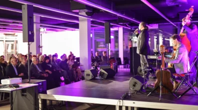 Na scenie w Mediatece trwa koncert podczas wydarzenia kulturalnego. Wokalista występuje przy mikrofonie, a obok niego muzycy grają na instrumentach, w tym na kontrabasie i gitarze. Przed sceną siedzi publiczność uważnie słuchająca występu. Przestrzeń oświetlona jest dekoracyjnym fioletowym światłem, które podkreśla koncertowy charakter wydarzenia.