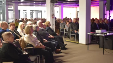 Publiczność siedzi w rzędach krzeseł i z uwagą obserwuje wydarzenie odbywające się przed nimi. W pierwszych rzędach widoczne są osoby w różnym wieku, a w tle znajduje się większa grupa uczestników spotkania. Obok ustawiony jest stolik z projektorem, a przestrzeń oświetlona jest światłem scenicznym w odcieniach fioletu i ciepłej bieli.