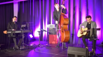 Na scenie występuje trzech muzyków podczas koncertu. Jeden z nich gra na instrumencie klawiszowym i śpiewa do mikrofonu, drugi gra na kontrabasie, a trzeci siedzi z gitarą akustyczną. Scena oświetlona jest fioletowym światłem, a wokół widoczny jest sprzęt nagłośnieniowy oraz instrumenty muzyczne.