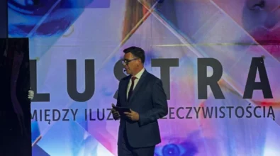 Zdjęcie przedstawia mężczyznę w okularach i garniturze. Stoi on na scenie, w tle widać grafikę przedstawiającą twarz w różnym ułożeniu oraz napis \