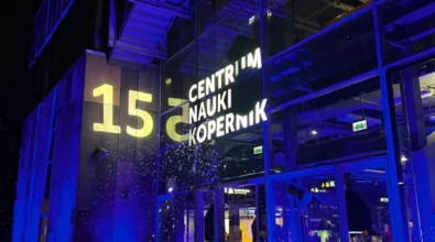 Zdjęcie przedstawiające wejście do Centrum Nauki Kopernik, co można wywnioskować po znajdującym się nad drzwiami napisem. Obok widać dużą liczbę 15, oznaczającą urodziny Centrum. Ściana budynku jest przeszklona, pada na nią granatowe światło. Zdjęcie: MBP.