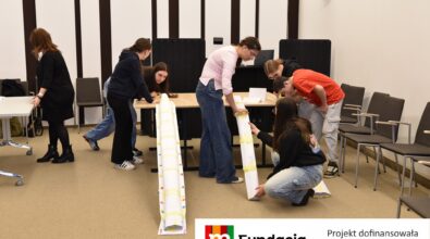 Grupa młodzieży konstruuje długie papierowe elementy przy stołach. Na dole logotyp Fundacji mBanku.