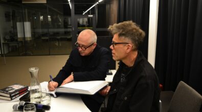 Dwie osoby siedzą przy stole; jedna pisze w dużej otwartej księdze.