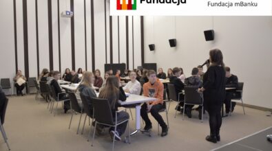 Uczestnicy konkursu słuchają kobiety mówiącej do mikrofonu. Na górze logotyp Fundacji mBanku.