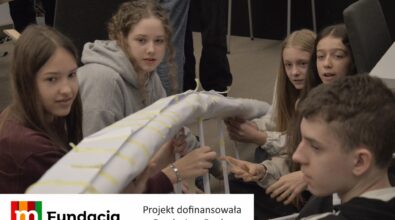 Młodzi uczestnicy analizują i modyfikują dużą papierową konstrukcję. Na dole logotyp Fundacji mBanku.