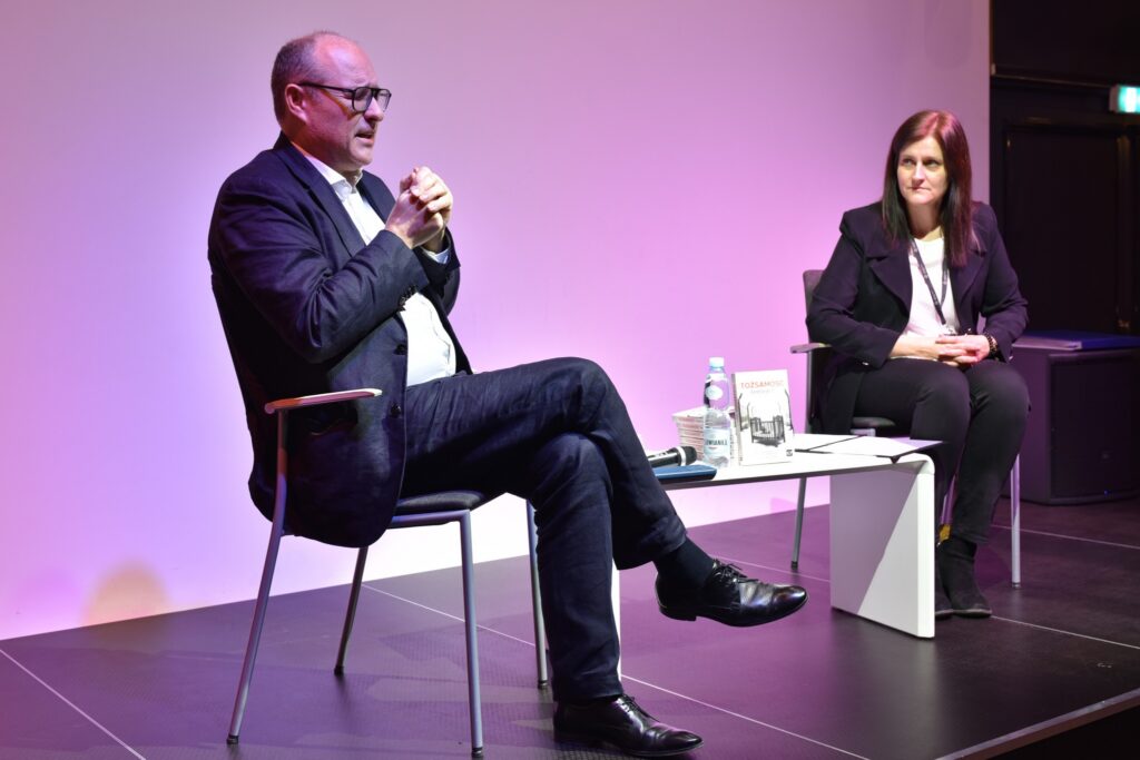 Błażej Torański i prowadząca spotkanie siedzą na scenie przy niskim białym stoliku.