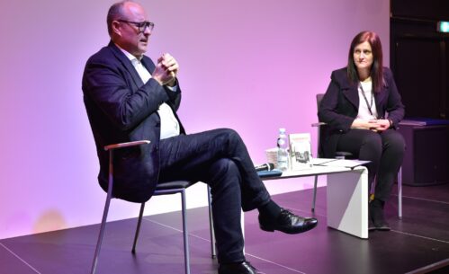 Błażej Torański i prowadząca spotkanie siedzą na scenie przy niskim białym stoliku.