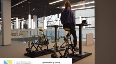 Osoba siedzi na rowerze biurowym przy stoliku, wytwarzając energię do ładowania urządzeń. Na dole zdjęcia logotypy.