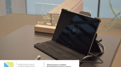 Tablet z widocznym stanem ładowania 80% obok statuetki „EkoTEKA” na stole w nowoczesnym wnętrzu. Na dole zdjęcia logotypy.