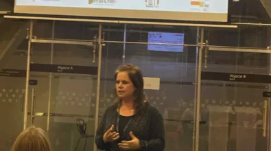 Zdjęcie przedstawia prezentującą jakiś temat osobę. U góry widać wyświetloną prezentację podsumowującą W tle widać szklane drzwi oraz ścianę. Z lewej strony siedzi kobieta, która patrzy na prezentację. Po środku stoi gestykulująca kobieta, która rozmawia z uczestnikami. Na prezentacji widoczne są logotypy miasta Piotrkowa Trybunalskiego, Miejskiej Biblioteki Publicznej im. Adama Próchnika oraz strefy SOWA. Zdjęcie zrobiono w Centrum Nauki Kopernik. Zdjęcie własnością MBP.