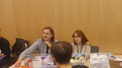 Zdjęcie przedstawia cztery osoby. Dwie osoby, kobiety, są w centrum zdjęcia. Obie mają okulary i siedzą frontem do oglądającego. Pokazują coś mężczyźnie w okularach, który siedzi po drugiej stronie stołu. Na stole widoczne są pudełka z różnymi elementami. Widoczne są klocki, piłki, tuby, zapakowane elementy, a także pomarańczowa konstrukcja z klocków. Jedna z kobiet trzyma biały element, który przykłada do konstrukcji. Z lewej strony widać ramię czwartej osoby oraz plecak. Z prawej strony widać pudełko i ubrania. W tle widoczna jest jasna ściana i szara wykładzina. Zdjęcie zrobiono w Centrum Nauki Kopernik. Zdjęcie własnością MBP.