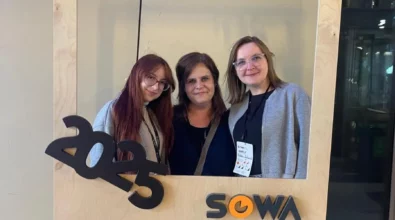 Zdjęcie przedstawia trzy kobiety. Dwie stojące z zewnątrz mają okulary. Osoba stojąca z lewej ma włosy w kolorze czerwonym. Po środku stoi szatynka, a z prawej strony blondynka. Przed osobami znajduje się zawieszona drewniana ramka. W górnym prawym rogu jest skośny napis Forum. W lewym dolnym rogu znajduje się rok 2025. W prawym dolnym rogu ramki jest napis SOWA. W tle widać pastelową ścianę oraz przeszkloną przestrzeń. Mogą to być drzwi. Zdjęcie zrobione zostało w Centrum Nauki Kopernik. Zdjęcie własnością MBP.