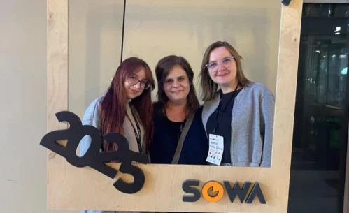 Zdjęcie przedstawia trzy kobiety. Dwie stojące z zewnątrz mają okulary. Osoba stojąca z lewej ma włosy w kolorze czerwonym. Po środku stoi szatynka, a z prawej strony blondynka. Przed osobami znajduje się zawieszona drewniana ramka. W górnym prawym rogu jest skośny napis Forum. W lewym dolnym rogu znajduje się rok 2025. W prawym dolnym rogu ramki jest napis SOWA. W tle widać pastelową ścianę oraz przeszkloną przestrzeń. Mogą to być drzwi. Zdjęcie zrobione zostało w Centrum Nauki Kopernik. Zdjęcie własnością MBP.