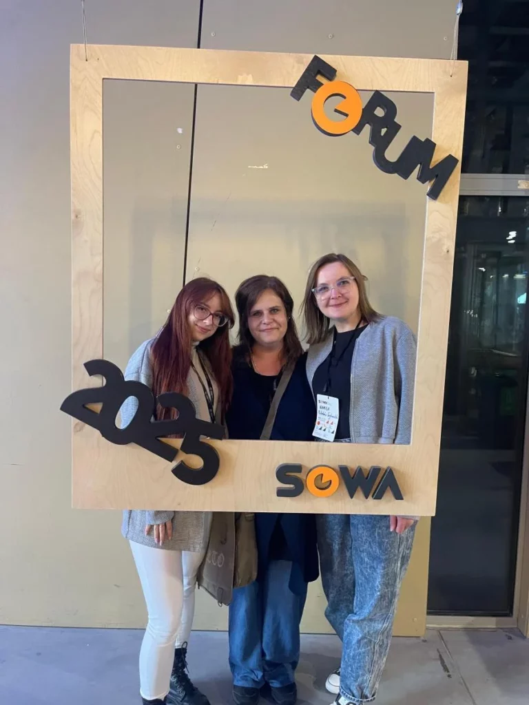 Zdjęcie przedstawia trzy kobiety. Dwie stojące z zewnątrz mają okulary. Osoba stojąca z lewej ma włosy w kolorze czerwonym. Po środku stoi szatynka, a z prawej strony blondynka. Przed osobami znajduje się zawieszona drewniana ramka. W górnym prawym rogu jest skośny napis Forum. W lewym dolnym rogu znajduje się rok 2025. W prawym dolnym rogu ramki jest napis SOWA. W tle widać pastelową ścianę oraz przeszkloną przestrzeń. Mogą to być drzwi. Zdjęcie zrobione zostało w Centrum Nauki Kopernik. Zdjęcie własnością MBP.
