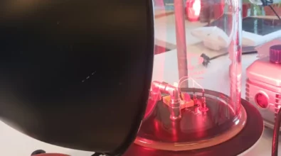 Zdjęcie przedstawia wnętrze laboratorium. Na pierwszym planie widoczna jest szklana kopuła i lampa z światem o czerwonym kolorze. Urządzenie jest podłączone przewodami. W tle widać okulary ochronne oraz inny sprzęt laboratoryjny. W środku kopuły widoczna jest elektronika. Pod spodem widoczne są podłączenia rurami. Wszystko znajduje się na blacie laboratoryjnym. Zdjęcie zrobiono w Centrum Nauki Kopernik. Zdjęcie własnością MBP.