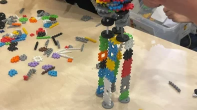 Zdjęcie przedstawia konstrukcję z kolorowych elementów o różnych kształtach. Konstrukcja wygląda jak niestabilna i krzywa wieża. Na jej wierzchołku znajduje się biały elementy przypominający kratkę. Konstrukcja stoi na jasnym blacie, prawdopodobnie stole. Dookoła widoczne są porozrzucane elementy kolorowe w różnym kształcie. Obok stołu stoją i siedzą cztery osoby. Widoczne są dłonie trzymające elementy. Zdjęcie zrobiono w Centrum Nauki Kopernik. Zdjęcie własnością MBP.