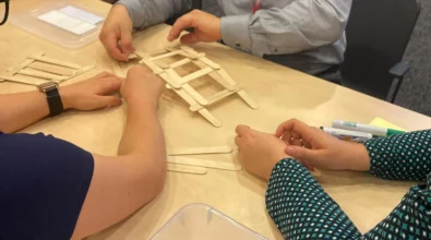 Zdjęcie przedstawia konstrukcję z drewnianych dużych patyczków do lodów. Konstrukcja przypomina most i jest umieszczona na jasnym blacie, prawdopodobnie stole. Przy nim widoczne są ręce czterech osób, prawdopodobnie jednego mężczyzny i trzech kobiet. Na jednej ręce widoczny jest zegarek. Na stole widoczne jest także pudełko plastikowe oraz flamastry w kolorze zielonym i czarnym. Zdjęcie zrobiono w Centrum Nauki Kopernik. Zdjęcie własnością MBP.