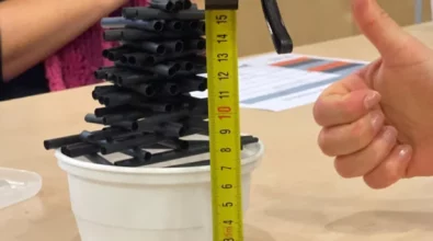 Zdjęcie prezentuje konstrukcję z białej plastikowej miski oraz czarnych słomek. Na pierwszym planie jest taśma miernicza z zaznaczonym pomiarem 15 cm, trzymającą ją dłoń kobiety oraz druga dłoń pokazująca gest w formie podniesionego kciuka. W tle widać kobietę z różowym szalikiem oraz kartkę z kolorowym nadrukiem. Konstrukcja znajduje się na jasnym blacie, prawdopodobnie stole. Zdjęcie zrobiono w Centrum Nauki Kopernik. Zdjęcie własnością MBP.