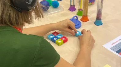 Zdjęcie przedstawia trzy kobiety siedzące przy stole. Jedna z kobiet ma na głowie słuchawki. Jednocześnie układa kolorowe klocki, na których widoczne są cyfry. Druga kobieta układa flamastry i długopis. Trzecia w ręku trzyma pomarańczową piłkę z wypustkami. Na środku stołu stoją plastikowe pojemniki z kolorowymi elementami w środku, prawdopodobnie tuby sensoryczne. Z lewej strony widocznych jest pięć różnokolorowych piłek o różnej strukturze. Z prawej strony widoczna jest kartka z napisem Majsternia oraz kolorowa karteczka. Zdjęcie zrobiono w Centrum Nauki Kopernik. Zdjęcie własnością MBP.