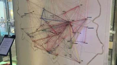 Zdjęcie przedstawia drewnianą mapę Polski. Zaznaczone są na niej miejscowości, w których znajdują się strefy SOWA. Miejsca połączone są kolorowymi sznurkami. Obok mapy stoi stojak z informacją, a także zielony taboret. Na taborecie leżą kolorowe sznurki i nożyczki. W tle widać przeszkloną przestrzeń. Prawdopodobnie jest to winda. Zdjęcie zrobione zostało w Centrum Nauki Kopernik. Zdjęcie własnością MBP.