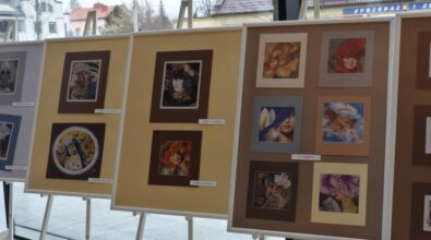 Na planszach ekspozycyjnych ustawionych na sztalugach zaprezentowane są oprawione prace artystyczne ułożone w kilku rzędach. Zdjęcie przedstawia fragment wystawy prezentowanej w przestrzeni Miejskiej Biblioteki Publicznej, widocznej na tle przeszklonej ściany.