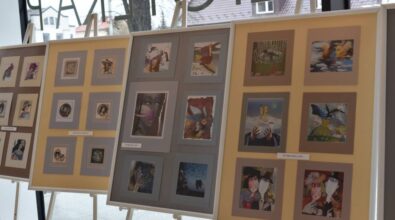 Na stojakach ekspozycyjnych prezentowane są plansze z fotografiami i pracami artystycznymi ułożonymi w równych rzędach. Zdjęcie przedstawia fragment wystawy artystycznej udostępnionej w przestrzeni Miejskiej Biblioteki Publicznej.