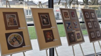 Na sztalugach ustawione są plansze ekspozycyjne z oprawionymi pracami artystycznymi, rozmieszczone w rzędzie przy przeszklonej ścianie. Zdjęcie przedstawia fragment wystawy prezentowanej w przestrzeni Miejskiej Biblioteki Publicznej, ukazującej różnorodne kompozycje i motywy artystyczne.