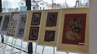 Na planszach ekspozycyjnych ustawionych na sztalugach zaprezentowane są oprawione prace artystyczne przedstawiające kobiece postacie i dekoracyjne kompozycje. Zdjęcie ukazuje fragment wystawy prezentowanej w przestrzeni Miejskiej Biblioteki Publicznej, z widocznym tłem przeszklonej elewacji i widokiem na zewnątrz budynku.