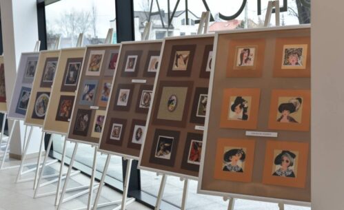 Na sztalugach ustawione są plansze ekspozycyjne z oprawionymi pracami artystycznymi przedstawiającymi portrety i ilustracje, rozmieszczone w kilku rzędach. Zdjęcie ukazuje fragment wystawy prezentowanej w przestrzeni Miejskiej Biblioteki Publicznej.