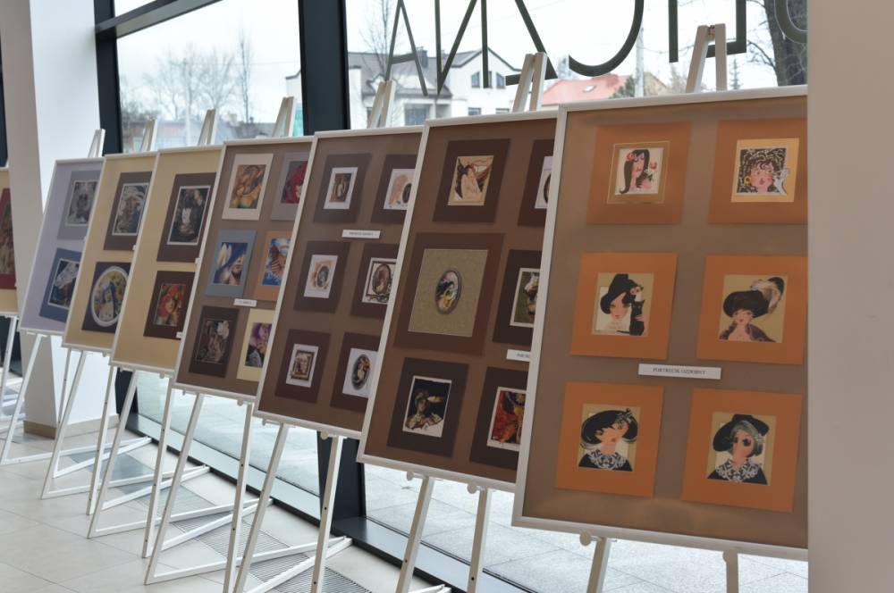 Na sztalugach ustawione są plansze ekspozycyjne z oprawionymi pracami artystycznymi przedstawiającymi portrety i ilustracje, rozmieszczone w kilku rzędach. Zdjęcie ukazuje fragment wystawy prezentowanej w przestrzeni Miejskiej Biblioteki Publicznej.