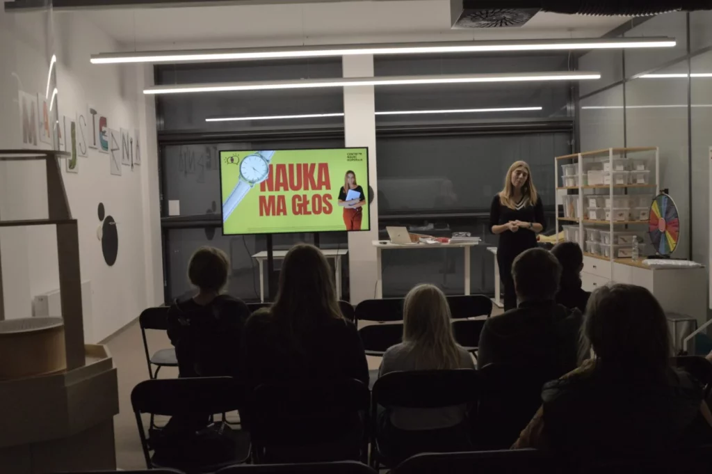 Zdjęcie przedstawiające spotkanie z kobietą w czarnym stroju. Obok niej stoi duży ekran, na którym wyświetlona jest grafika: na żółtym tle widać czerwony napis \"Nauka ma głos\". Na literze N leży biały zegarek na rękę z tarczą. Po prawej stronie grafiki widać zdjęcie kobiety prowadzącej spotkanie. Obok ekranu widać stół, na którym znajduje się laptop oraz inne elementy. Po prawej stronie zdjęcia widać dużą półkę, na której stoją plastikowe pudełka. Po lewej stronie, na ścianie wisi napis \"Majsternia\", wykonany z grafik przedstawiających narzędzia. Zdjęcie: MBP.