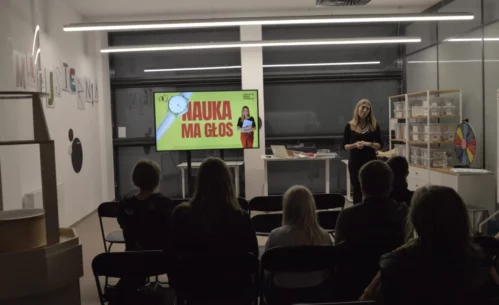Zdjęcie przedstawiające spotkanie z kobietą w czarnym stroju. Obok niej stoi duży ekran, na którym wyświetlona jest grafika: na żółtym tle widać czerwony napis \"Nauka ma głos\". Na literze N leży biały zegarek na rękę z tarczą. Po prawej stronie grafiki widać zdjęcie kobiety prowadzącej spotkanie. Obok ekranu widać stół, na którym znajduje się laptop oraz inne elementy. Po prawej stronie zdjęcia widać dużą półkę, na której stoją plastikowe pudełka. Po lewej stronie, na ścianie wisi napis \"Majsternia\", wykonany z grafik przedstawiających narzędzia. Zdjęcie: MBP.