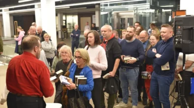 Czytelnicy ustawili się w kolejce, trzymając w rękach książki i czekając na autograf. Andrzej Pilipiuk stoi przy stoliku i rozmawia z uczestnikami spotkania, podpisując egzemplarze swoich książek. Wokół zgromadziła się grupa osób zainteresowanych rozmową z autorem oraz zdobyciem autografu. Wydarzenie odbywa się w przestrzeni Mediateki, gdzie uczestnicy mają możliwość bezpośredniego spotkania z pisarzem.