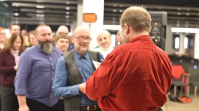 Andrzej Pilipiuk stoi tyłem do aparatu i podpisuje książkę jednemu z czytelników. Przed autorem ustawiła się kolejka osób trzymających egzemplarze książek i czekających na autograf. Najbliżej stoi mężczyzna, który podaje książkę do podpisania. W tle widać kolejnych uczestników spotkania autorskiego oczekujących na rozmowę z pisarzem.