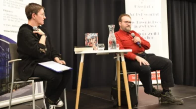 Prowadząca spotkanie siedzi na krześle i trzyma w ręku mikrofon oraz notatki. Obok przy stoliku siedzi Andrzej Pilipiuk, który odpowiada na pytania, trzymając mikrofon. Na stoliku znajdują się książki autora, karafka z wodą oraz szklanka. W tle widoczne są roll-upy organizatorów wydarzenia.