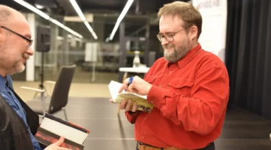 SAndrzej Pilipiuk podpisuje książkę jednemu z czytelników podczas spotkania autorskiego. Autor trzyma w dłoni długopis i składa autograf na egzemplarzu książki. Obok stoi uczestnik wydarzenia, który trzyma kolejną książkę przeznaczoną do podpisania. Spotkanie odbywa się w przestrzeni Mediateki, gdzie czytelnicy mają możliwość bezpośredniej rozmowy z pisarzem i zdobycia autografu.